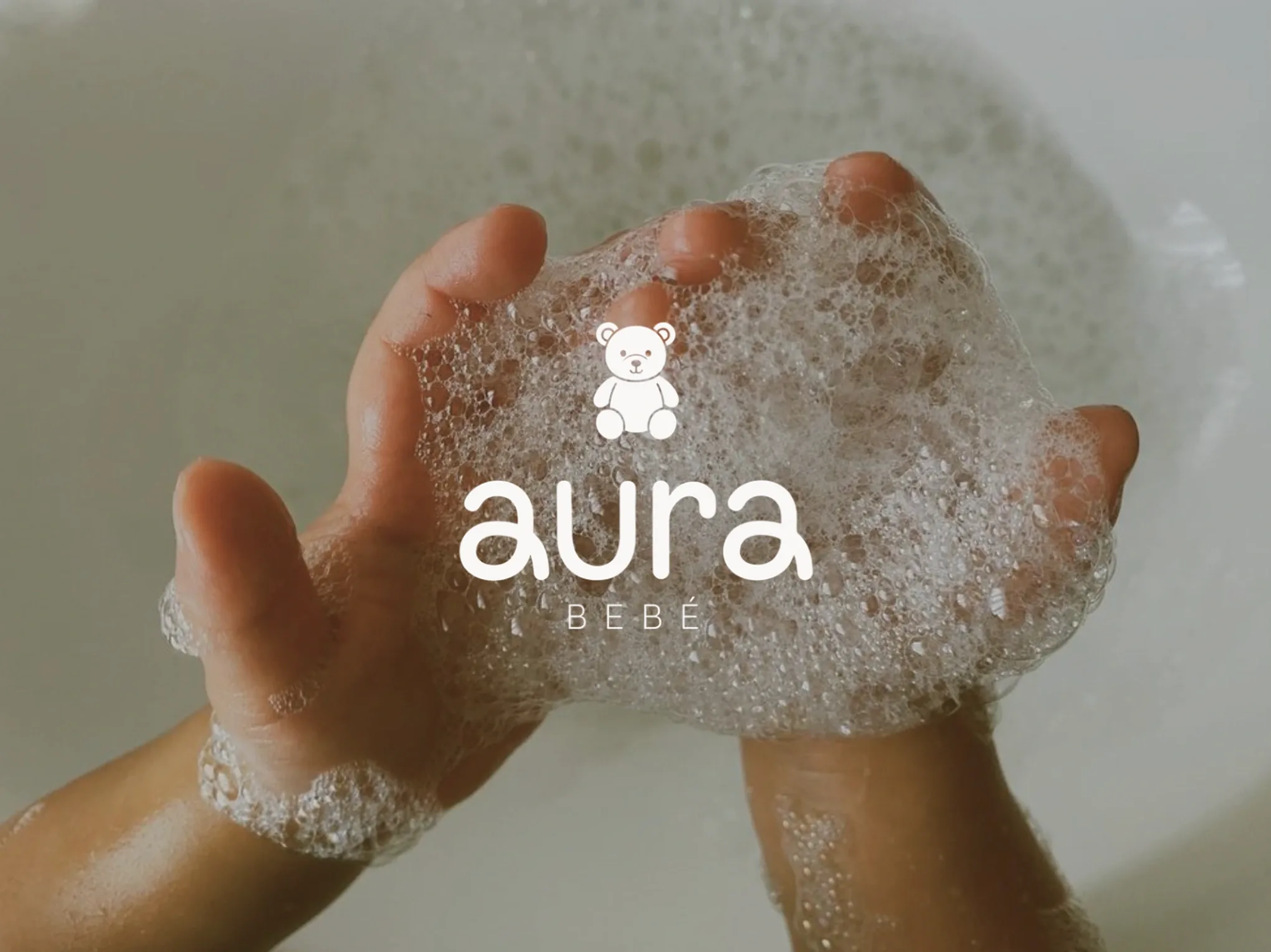 Logotipo de Aura Bebé con su osito sobre la fotografía de las manos de un bebé entre espuma