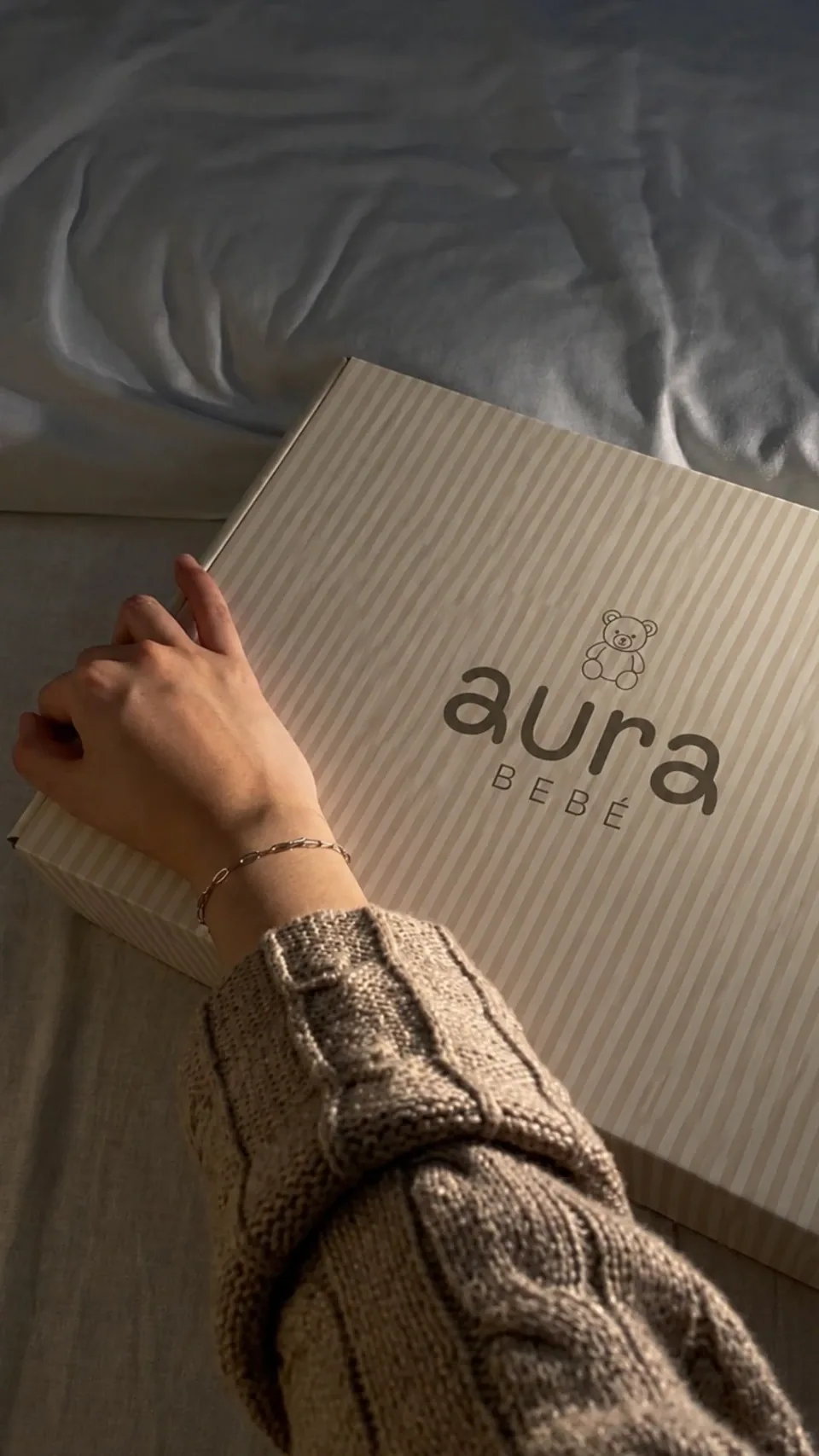 Caja de regalo beige con el logotipo de Aura Bebé, abierta por unas manos sobre una cama