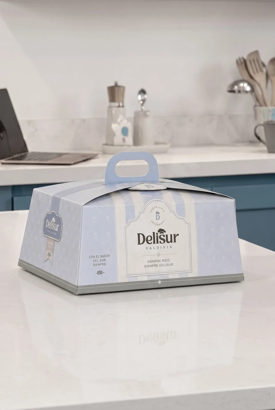 Caja de torta a rayas celestes con asa y el logotipo de Delisur, sobre un mesón de cocina